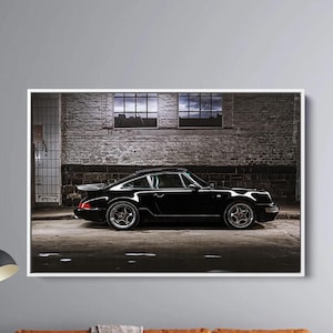 Op de afbeelding: Een ingelijste print met een zwarte sportwagen met een spoiler en zilveren velgen. De auto staat op straat, met een bakstenen muur en twee ramen op de achtergrond. De print is wit omlijst.