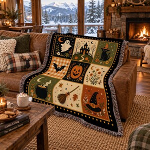 May include: A woven throw blanket featuring a grid of nine Halloween-themed illustrations, including a ghost, a haunted house, a black cat, a bat, a jack-o'-lantern, a cauldron, a broomstick, a witch hat, and floral motifs in orange, black, and green.