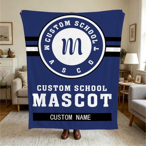 Op de afbeelding: Een blauwe plaid met een centraal rond embleem met de letter M, omringd door de tekst CUSTOM SCHOOL MASCO. Onder het embleem staan de woorden CUSTOM SCHOOL MASCOT in het wit, gevolgd door een zwarte balk met het opschrift CUSTOM NAME.