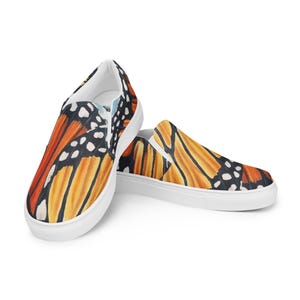 May include: A pair of slip-on shoes featuring a vibrant monarch butterfly wing design. The shoes showcase orange, black, and white patterns on a white sole. The design wraps around the top and sides of the shoe.
