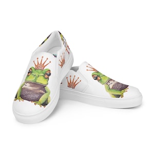 May include: White slip-on shoes featuring a watercolor illustration of a green frog wearing a crown. The frog has a brown belly and is set against a white background. The text "NO KINGS" is printed on the side of the shoe.