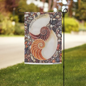 Könnte beinhalten: Eine Gartenflagge mit einem Nautilus-Muschel-Design und dem Schriftzug "Diversity is Beautiful". Die Flagge hat einen Hintergrund aus verschiedenfarbigen Kieselsteinen und wird auf einem schwarzen Metallständer in einem grasbewachsenen Garten gezeigt.