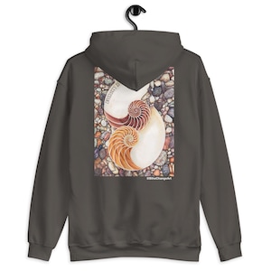 Puede incluir: Sudadera con capucha gris oscuro con un diseño de concha de nautilus colorido en la espalda. La obra de arte presenta una ilustración detallada de una concha de nautilus en tonos marrones y naranjas, sobre un fondo de guijarros. El texto "Diversity Is" está impreso en el lado izquierdo.