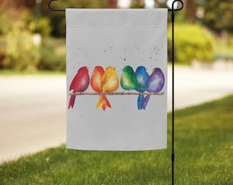 May include: A garden flag featuring a watercolor painting of six colorful birds perched on a branch. The birds are in shades of red, orange, yellow, green, blue, and purple. The background is light gray with colorful paint splatters.
