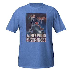 May include: Blue t-shirt featuring a graphic of a person as a puppet with the text "WHO PULLS THE STRINGS?". The puppet is controlled by two hands. The shirt is made of a soft material.