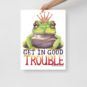 May include: A watercolour print of a green frog wearing a gold crown with the words "NO KINGS." Below the frog, the words "GET IN GOOD TROUBLE" are written in bold, colourful letters. The artist's signature is at the bottom.