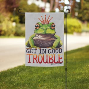 May include: A garden flag featuring a green frog wearing a crown with the words "NO KINGS." The flag also displays the text "GET IN GOOD TROUBLE" in orange and red. The flag is hanging on a black metal pole in a grassy yard.