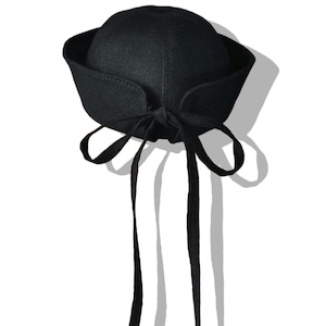 Puede incluir: Un sombrero negro con una copa redondeada y un ala curvada. El sombrero tiene largas cintas negras atadas con un lazo en la parte trasera. El sombrero está sobre un fondo blanco.