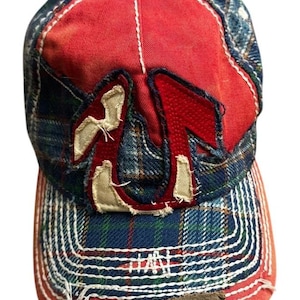 May include: A baseball cap featuring a red and blue plaid pattern with distressed edges. The front panel displays a large, red, horseshoe-shaped applique with frayed cream-colored fabric edges. The curved brim has thick white stitching and a brown patch.
