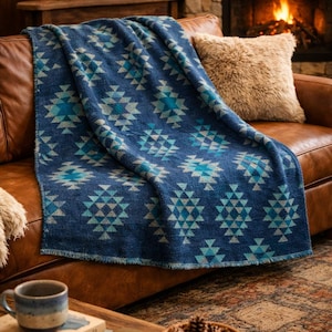 Op de afbeelding: Een blauwe sprei met een geometrisch patroon van turquoise en lichtblauwe driehoeken over een bruine leren bank. Een zacht beige kussen ligt op de bank. Een brandende open haard en een lantaarn staan op de achtergrond.