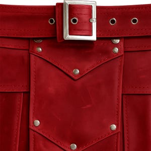 Op de afbeelding: Close-up van een rood leren kledingstuk met een zilveren gesp en klinknagels. De riem heeft een rechthoekige zilveren gesp en diverse oogjes. Het kledingstuk heeft een zak met decoratieve klinknagels.