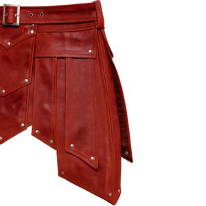 Op de afbeelding: Een rode leren kilt met een riem en gespsluiting. De kilt heeft decoratieve zilveren klinknagels en meerdere zakken. Het ontwerp omvat schuine panelen en een gestructureerd silhouet.