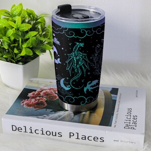 May include: Stainless steel travel mug with a black background and a teal dragon design. The design includes clouds, stars, and crescent moons. The lid is black with a sliding closure.