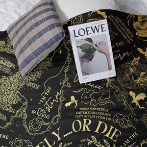 May include: A black blanket with a gold map design and text, including "I AM THE STORM" and "OR DIE." A "LOEWE" magazine and a plaid pillow rest on the blanket. The blanket's design features a map-like layout with various place names.
