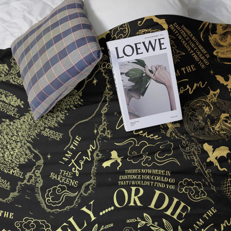 May include: A black blanket with a gold map design and text, including "I AM THE STORM" and "OR DIE." A "LOEWE" magazine and a plaid pillow rest on the blanket. The blanket's design features a map-like layout with various place names.