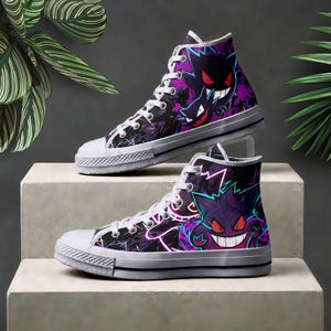 Puede incluir: Un par de zapatillas altas con un estampado gráfico en negro y morado de personajes Gengar con ojos rojos y dientes blancos. El calzado tiene cordones blancos, punteras de goma blanca y suelas de goma blanca con una fina línea negra.