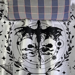 May include: A white blanket with a black dragon design, surrounded by a circular pattern of celestial elements and foliage. A blue, gray, and red plaid pillow rests at the top. The blanket is likely for a bed or couch.