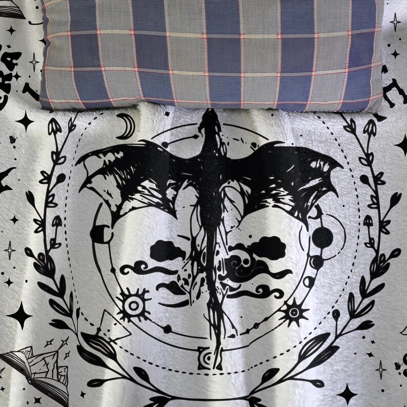 May include: A white blanket with a black dragon design, surrounded by a circular pattern of celestial elements and foliage. A blue, gray, and red plaid pillow rests at the top. The blanket is likely for a bed or couch.