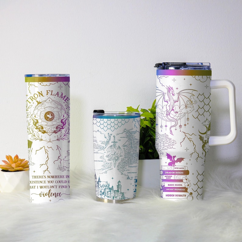 May include: Three white insulated tumblers with fantasy-themed designs. One features a dragon and the text "DRAGON RIDERS". Another has a map design, and the third has the text "IRON FLAME" and "violence". Each tumbler has a different lid and handle.