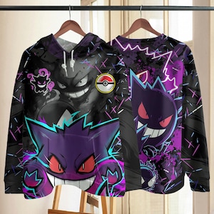 Puede incluir: Una sudadera con capucha negra que presenta un estampado vibrante de cuerpo entero del Pokemon morado Gengar con ojos rojos y una amplia sonrisa. El diseño incluye acentos de rayos azul neón y rosa, un icono de Pokeball y fondo oscuro.