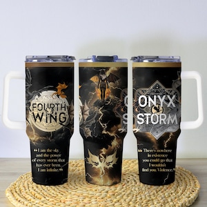 May include: Three black insulated tumblers with white handles and lids. Each tumbler features a different design with gold accents and text. The text includes "FOURTH WING" and "ONYX STORM" with quotes. The tumblers are on a woven mat.
