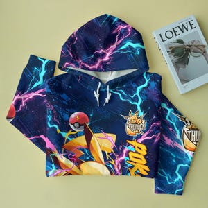 May include: A dark blue hoodie featuring a vibrant pattern of pink and cyan lightning bolts. The design includes a yellow character, a red and white ball, and the text "POK&Eacute;MON" and "THUNDER" in bold yellow letters across the fabric surface.