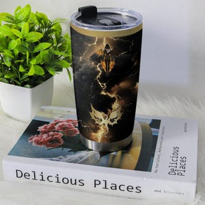 May include: A stainless steel travel mug featuring a dramatic design of a winged figure against a stormy sky with lightning. The mug has a black lid and a gold band near the top.