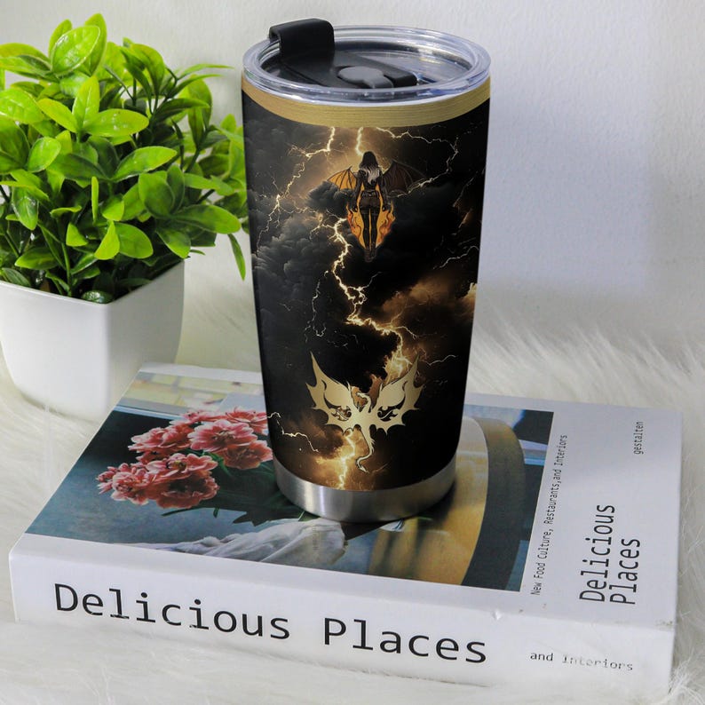 May include: A stainless steel travel mug featuring a dramatic design of a winged figure against a stormy sky with lightning. The mug has a black lid and a gold band near the top.