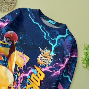 May include: A long-sleeve sweatshirt featuring a dark blue background with bright cyan and magenta lightning patterns. The design includes a yellow character, a red and white ball, and a logo with the text THUNDER and a character illustration.