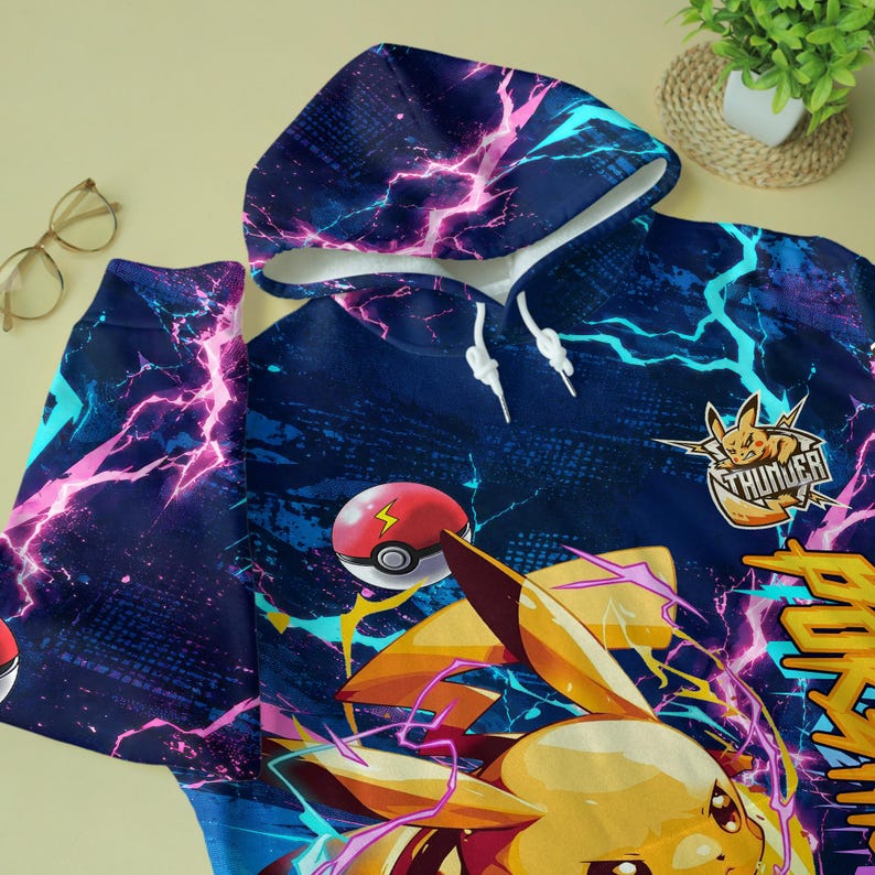 May include: A dark blue hoodie featuring a vibrant graphic print of a yellow electric creature, a red and white ball, and bright pink and cyan lightning bolts. The design includes the text "THUNDER" and "POKEMON" in bold yellow lettering.