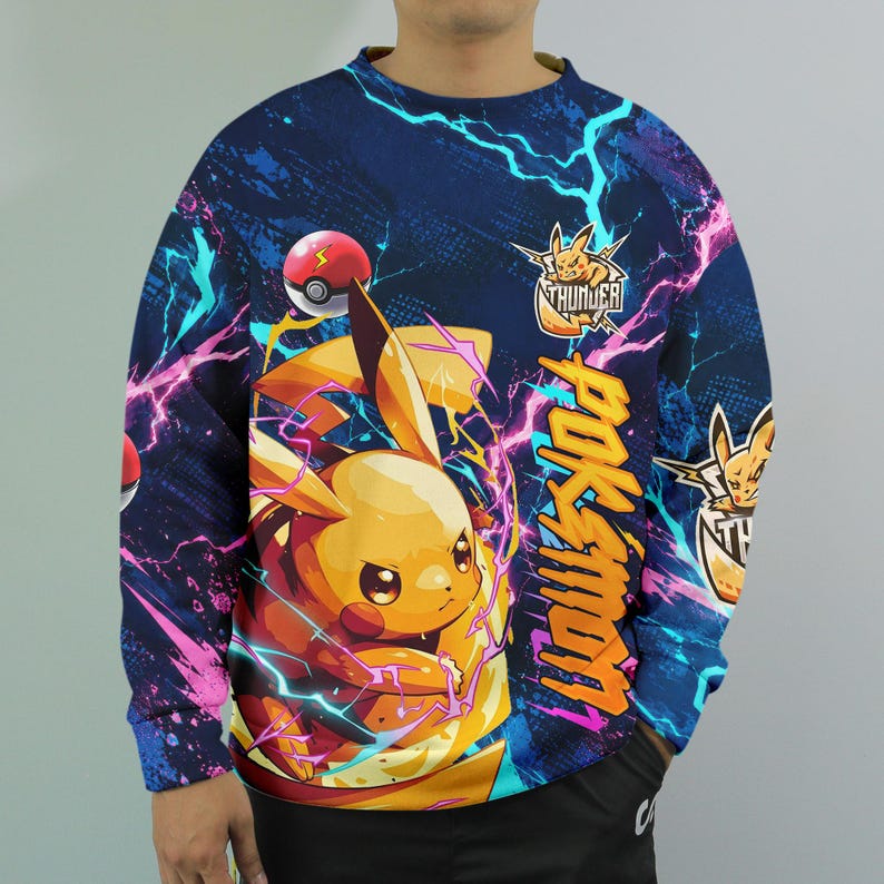 May include: A long-sleeve sweatshirt featuring a vibrant graphic of a yellow electric mouse character surrounded by pink and blue lightning bolts. The design includes a red and white ball, the text "POKEMON" in orange, and a "THUNDER" logo.
