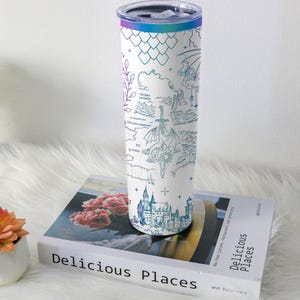 May include: A white stainless steel tumbler with a fantasy map design in shades of blue and teal. The map includes illustrations of a dragon, castle, and other fantastical elements. The tumbler has a gradient lid and is resting on a book titled "Delicious Places."