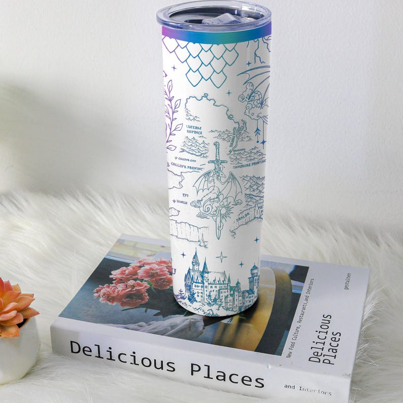 May include: A white stainless steel tumbler with a fantasy map design in shades of blue and teal. The map includes illustrations of a dragon, castle, and other fantastical elements. The tumbler has a gradient lid and is resting on a book titled "Delicious Places."