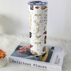 May include: White insulated tumbler with a gold dragon and book pattern. The tumbler has a silver lid and the text "PLACE YOU COULD GO". The tumbler is sitting on a book titled "Delicious Places".
