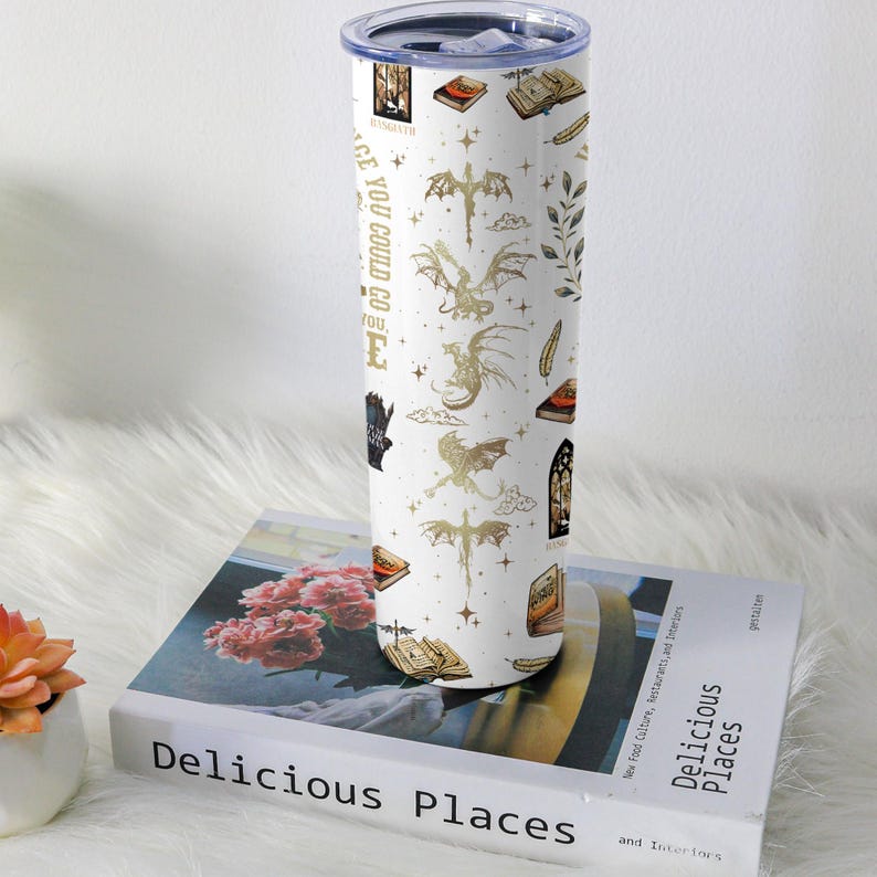 May include: White insulated tumbler with a gold dragon and book pattern. The tumbler has a silver lid and the text "PLACE YOU COULD GO". The tumbler is sitting on a book titled "Delicious Places".