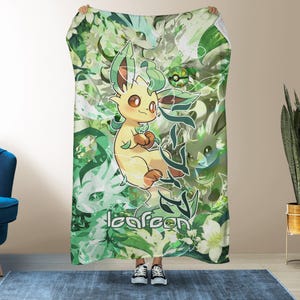 May include: A rectangular throw blanket featuring a cream-colored Leafeon character surrounded by various green foliage, white flowers, and abstract leaf patterns. The text "leafeon" is printed in white, along with Japanese characters and a small Poké Ball icon.