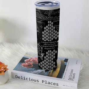 May include: A stainless steel travel tumbler with a black background featuring a dragon scale design and text. The text reads "A dragon without its rider is a tragedy. A dragon without their dragon is dead." The tumbler is cylindrical and has a silver lid.
