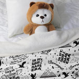 May include: A brown teddy bear wearing a bear-themed hood is nestled under a white blanket. The blanket features a black and white design with text that reads "In My Sarah J. Maas Era" and other phrases and illustrations.