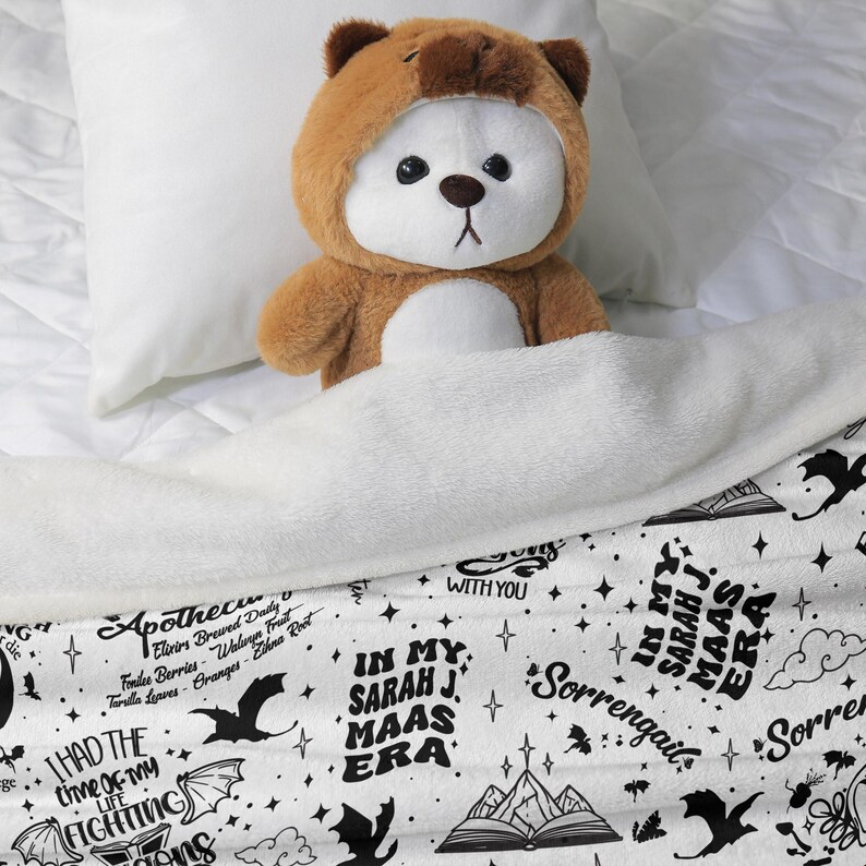 May include: A brown teddy bear wearing a bear-themed hood is nestled under a white blanket. The blanket features a black and white design with text that reads "In My Sarah J. Maas Era" and other phrases and illustrations.