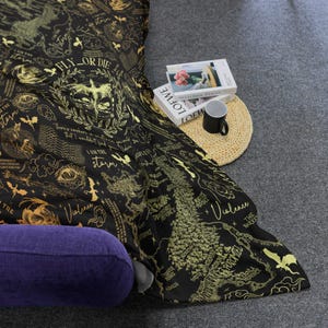 May include: Black and gold patterned blanket with text and illustrations. The blanket features phrases like "FLY OR DIE" and "I AM THE STORM", along with a map design and other graphics. A purple pillow is visible.