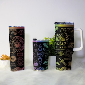 May include: Three black travel mugs with vibrant gradient designs. The mugs display text and illustrations, including "Rhysand's Tattoo Parlor," "Elaris City of Starlight," and "Hello Feyre darling." The designs incorporate moons, stars, and mountain scenes.