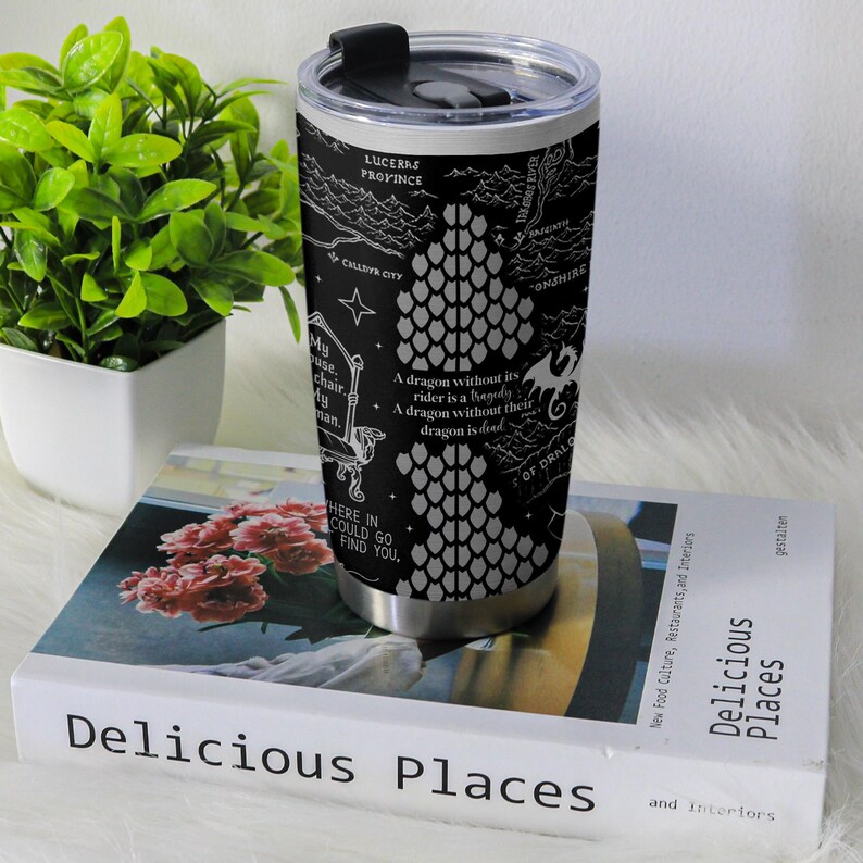 May include: A black travel mug with a silver base and a black lid. The mug features a white dragon scale design and text, including "A dragon without its rider is a tragedy." The mug is on a book titled "Delicious Places."