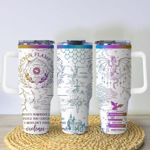 May include: Three white travel mugs with colorful lids and handles. Each mug features a different fantasy-themed design with text. One mug has a dragon and the words "Dragon Riders". The mugs are on a woven mat.