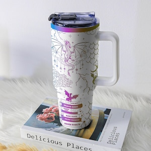 May include: White travel mug with a handle, featuring a dragon design in purple, pink, and gold. The mug includes text that reads "DRAGON RIDERS", "MAGIC SCHOOL", and "HIDDEN ROMALTY". The lid and rim have a rainbow gradient.