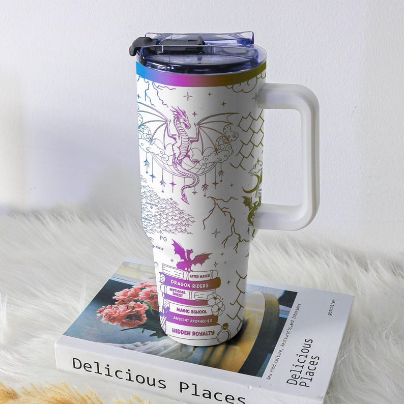 May include: White travel mug with a handle, featuring a dragon design in purple, pink, and gold. The mug includes text that reads "DRAGON RIDERS", "MAGIC SCHOOL", and "HIDDEN ROMALTY". The lid and rim have a rainbow gradient.
