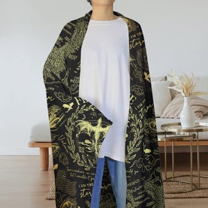May include: A black and gold patterned shawl with the text "I AM THE storm" and other phrases. The shawl features illustrations of dragons, stars, and other celestial elements. The person is wearing a white shirt and blue jeans.