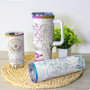 May include: White insulated tumblers with fantasy-themed designs. One tumbler features a dragon, another a map, and a third has the text "Iron Flame" and "violence". Each tumbler has a colorful rim.