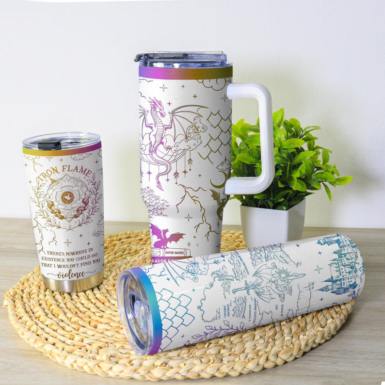 May include: White insulated tumblers with fantasy-themed designs. One tumbler features a dragon, another a map, and a third has the text "Iron Flame" and "violence". Each tumbler has a colorful rim.