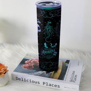 May include: A black insulated tumbler with a dragon and cloud design, and the text "ANDARNA WE DO NOT EAT OUR ALLIES". The tumbler has a teal and purple color scheme and a silver lid. It is sitting on a book.