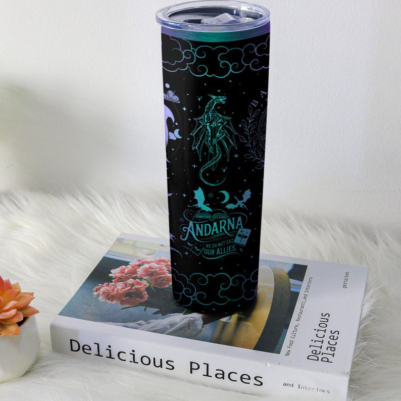 May include: A black insulated tumbler with a dragon and cloud design, and the text "ANDARNA WE DO NOT EAT OUR ALLIES". The tumbler has a teal and purple color scheme and a silver lid. It is sitting on a book.